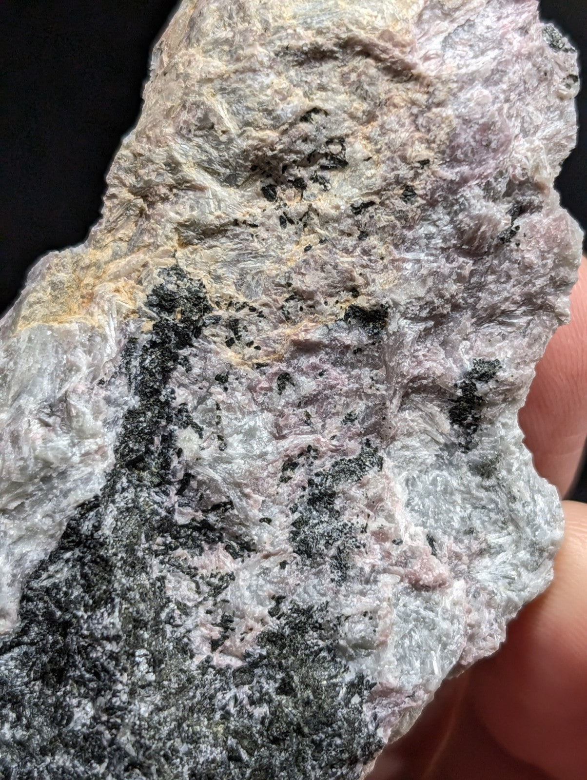 A rough, irregularly shaped mineral specimen with a mottled surface of gray, white, and subtle pinkish hues, featuring dark black streaks and speckles.