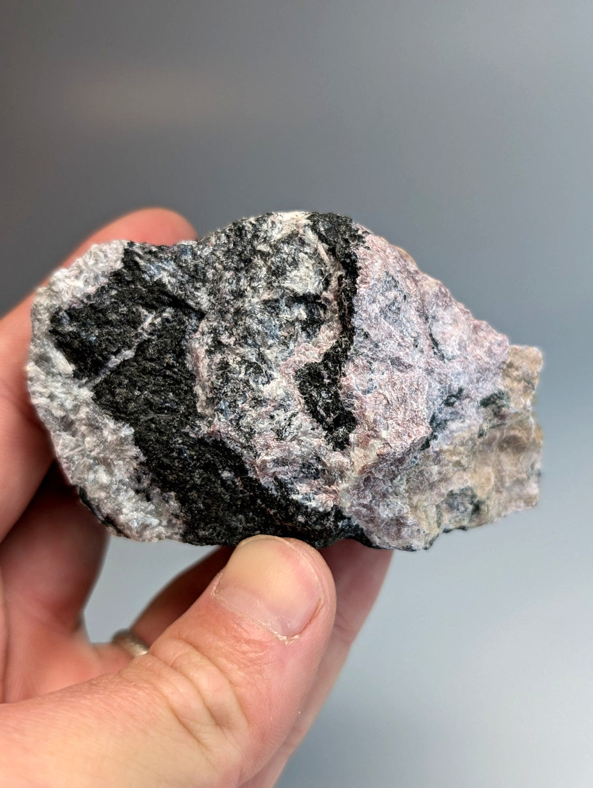 A hand holds a rough miserite and wollastonite rock with black gray and pink swirling patterns from Arkansas USA