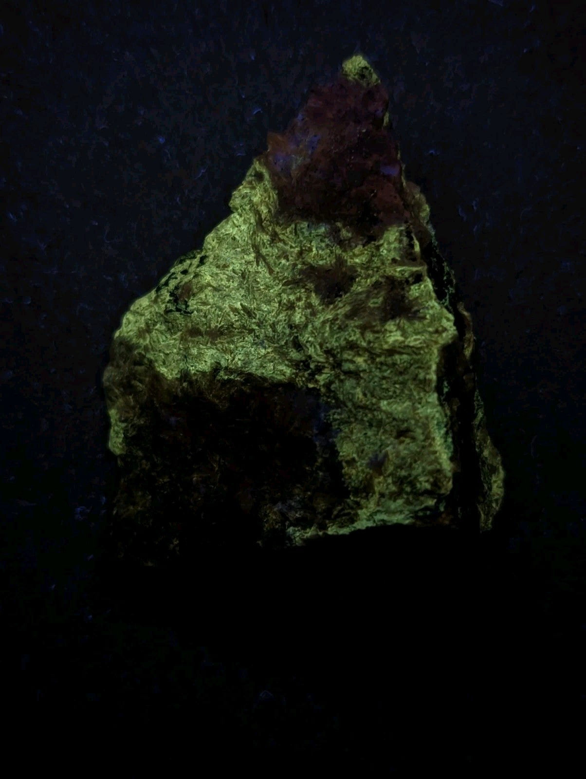 A rough, irregularly shaped mineral specimen with a vibrant greenish-yellow surface and a small patch of reddish-purple at its peak.