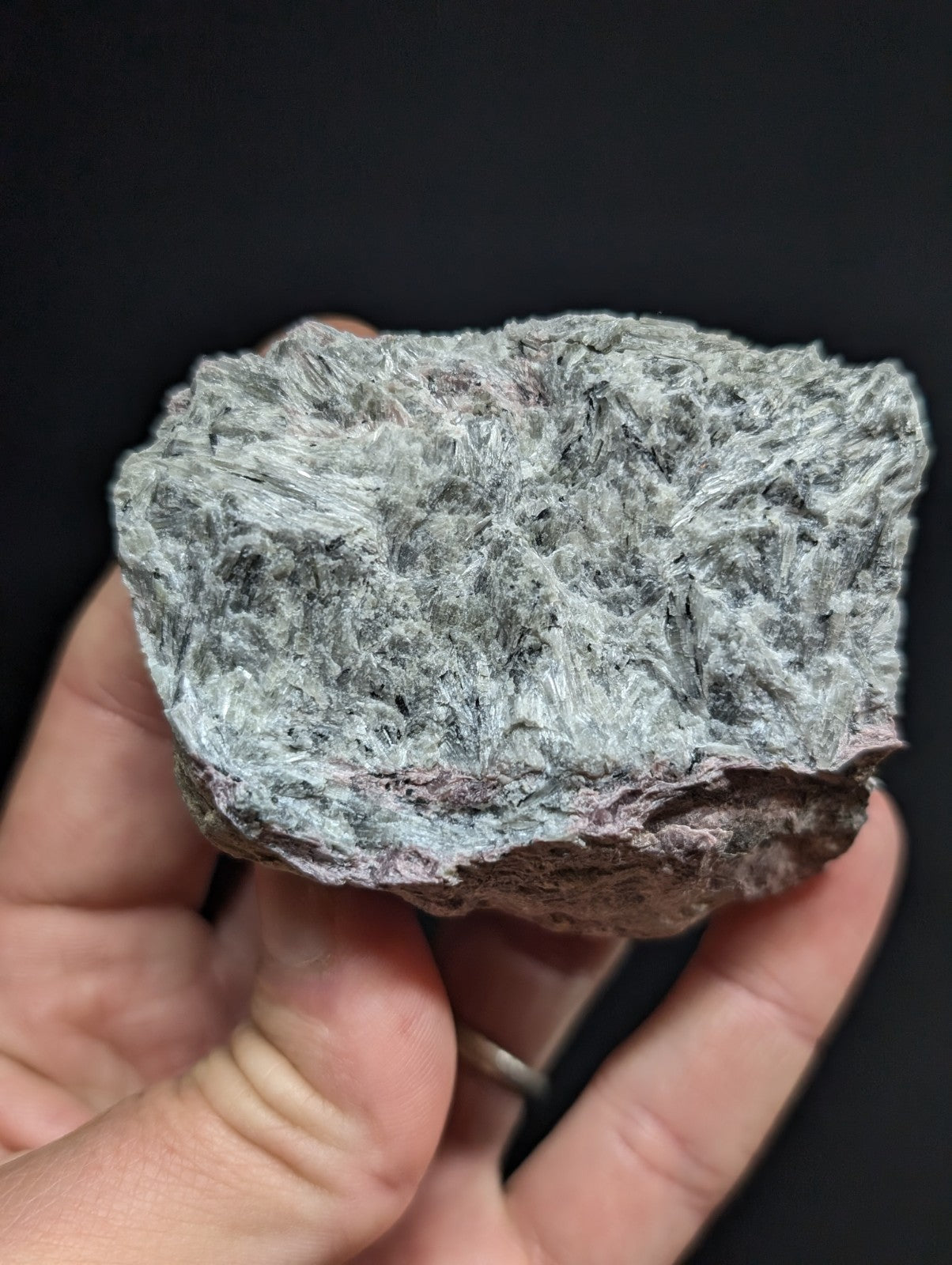 A hand holds a rough, irregularly shaped piece of grayish-white fibrous mineral with subtle reddish-brown undertones and textured surface.