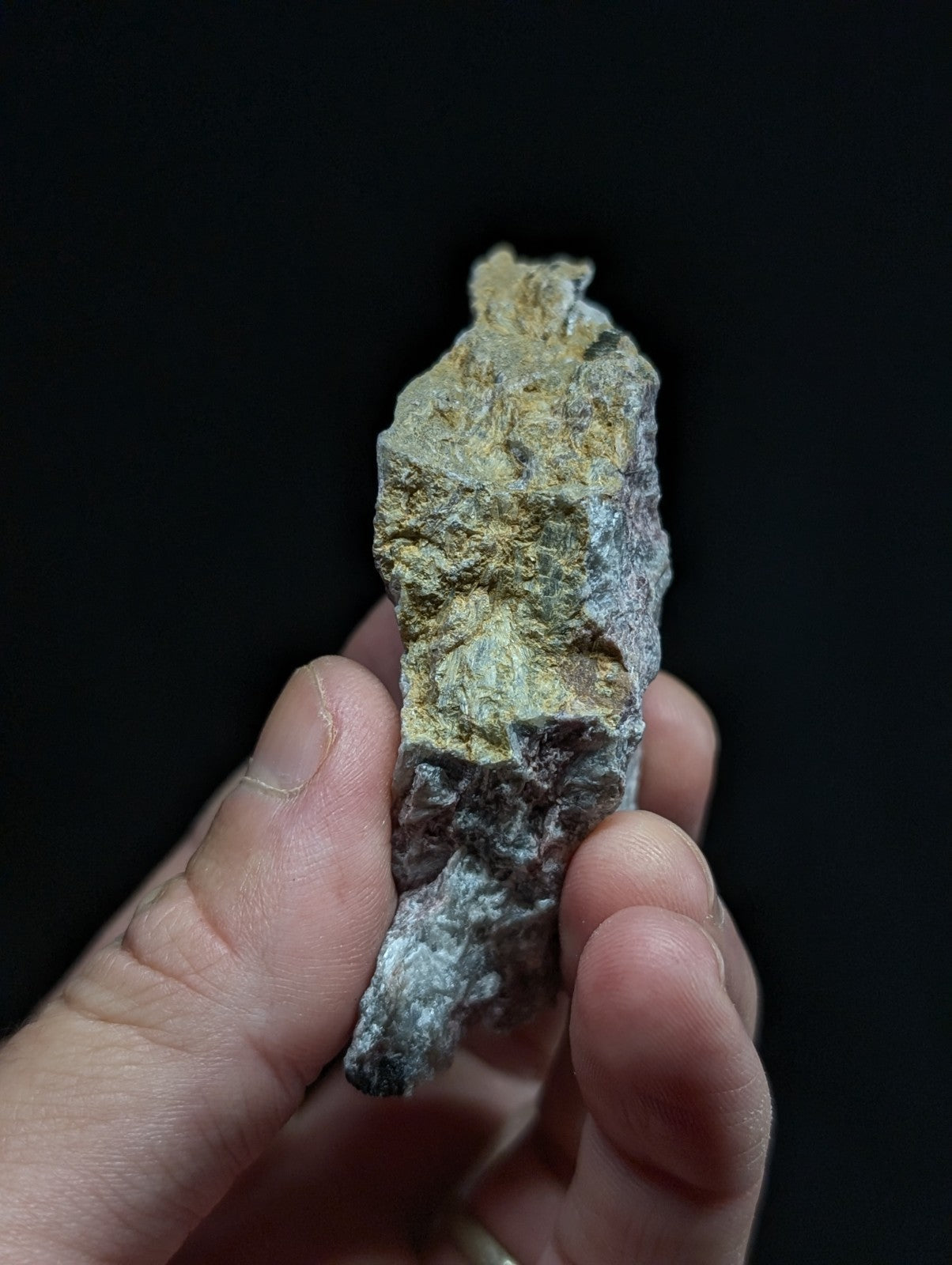 A rough, irregularly shaped mineral specimen with a mottled surface of gray, greenish-yellow, and dark patches.