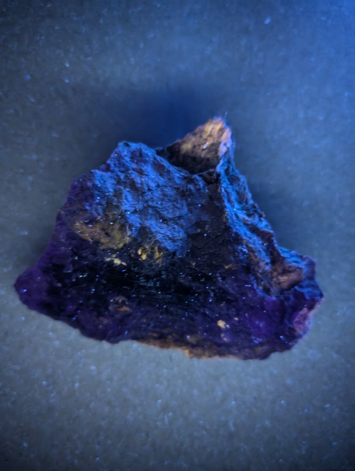 A rugged, irregularly shaped purple mineral specimen with a dark, textured surface and patches of golden-yellow streaks.