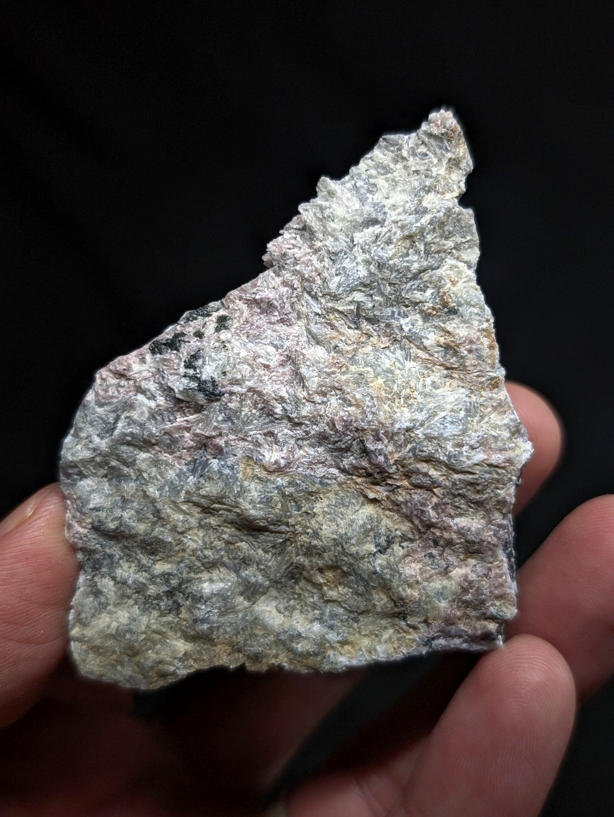 A rough, irregularly shaped piece of grayish-white rock with subtle hints of green and brown, held gently in a hand against a dark background.