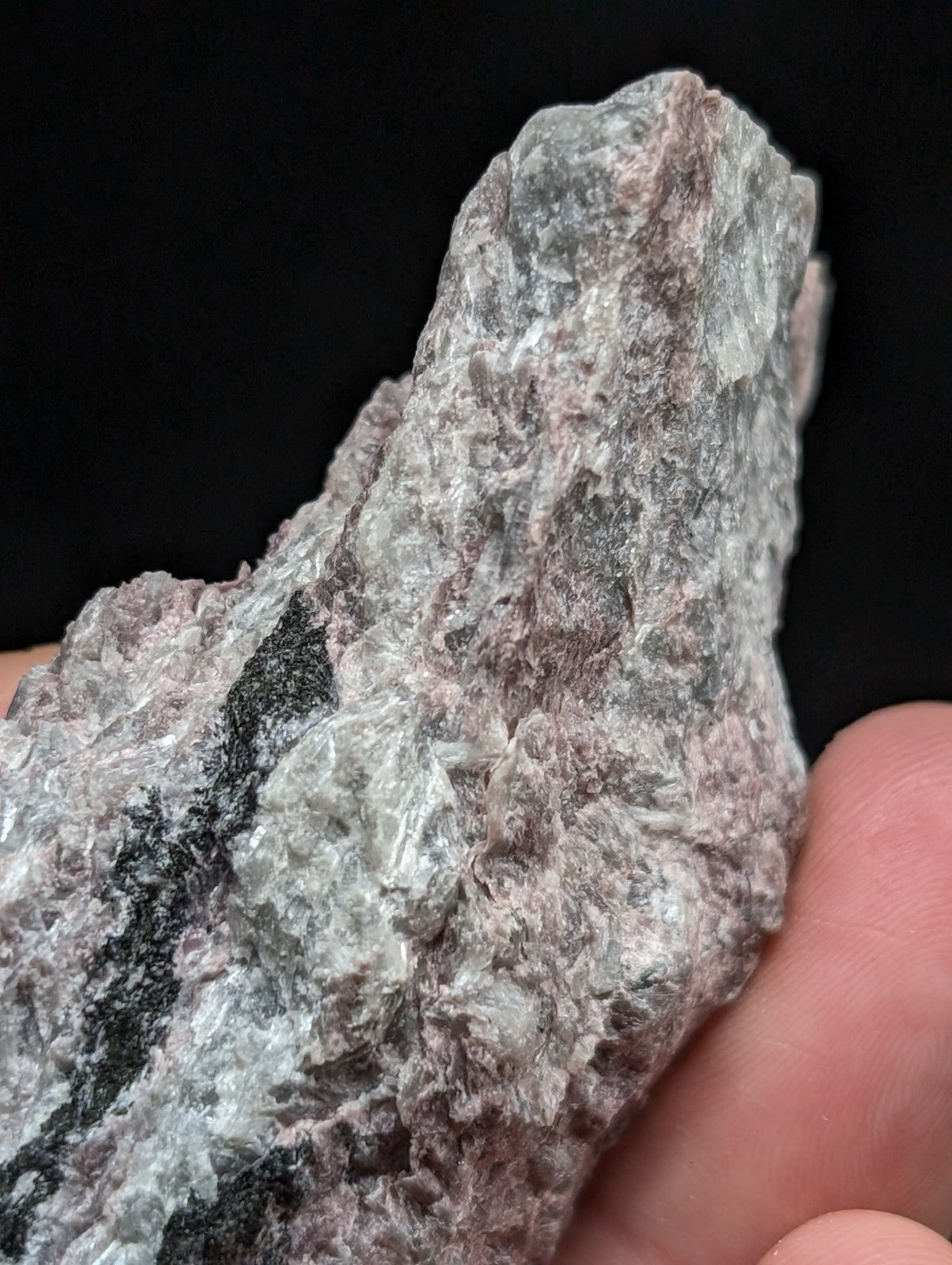 A rough, irregularly shaped mineral specimen with a mottled surface of gray, pinkish hues and dark streaks.