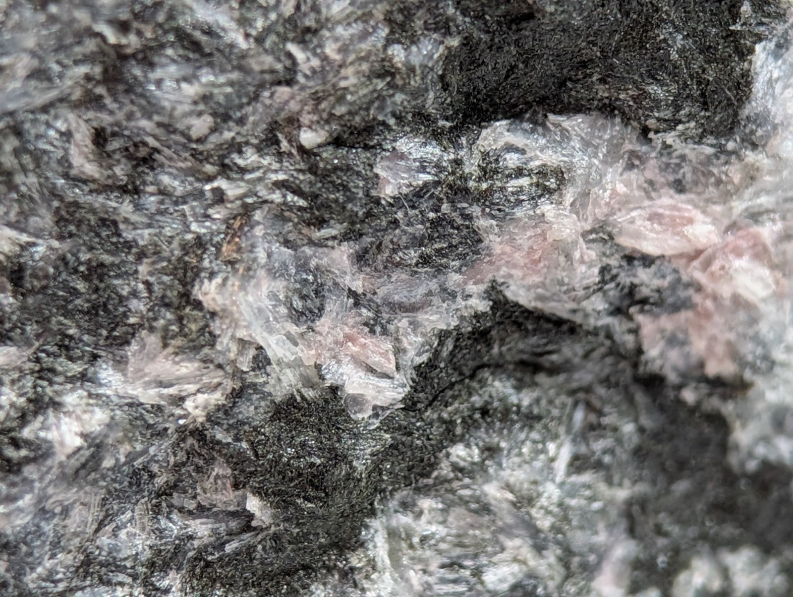 A rugged, speckled rock with a mix of dark gray and black tones interspersed with translucent white crystals and subtle pinkish mineral inclusions.