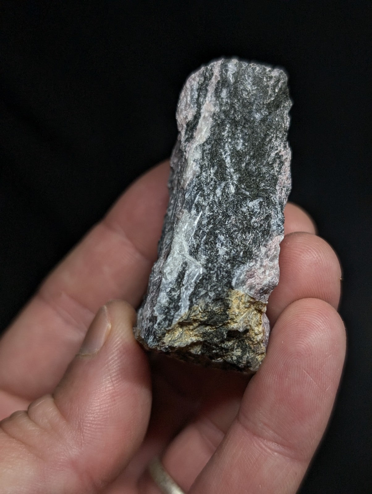 A rough, irregularly shaped stone with a mottled gray and white surface, featuring subtle veining and a small patch of yellowish mineral at its base.