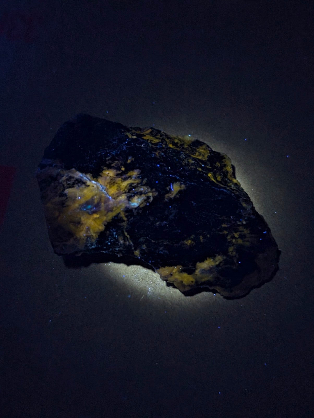 A rugged, irregularly shaped piece of dark mineral with patches of vibrant yellow and iridescent blue-green flecks.