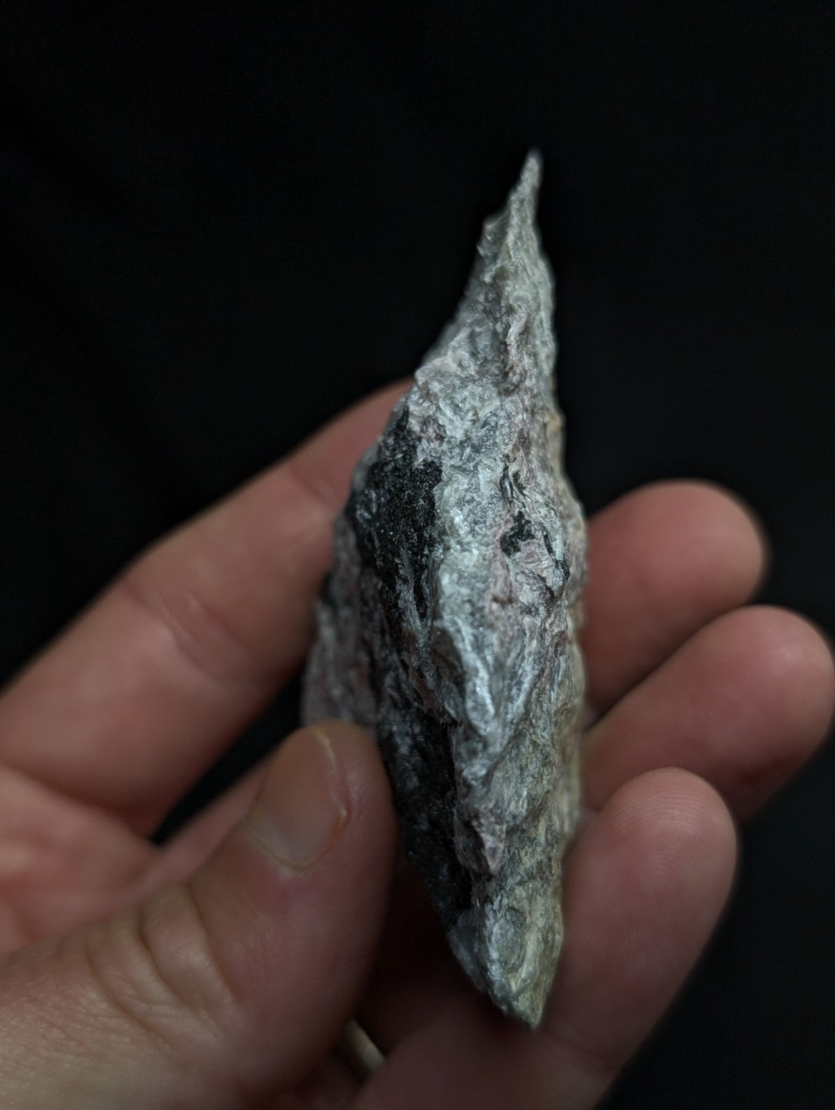 A hand holds a rough, irregularly shaped gray rock with dark patches and a pointed tip.