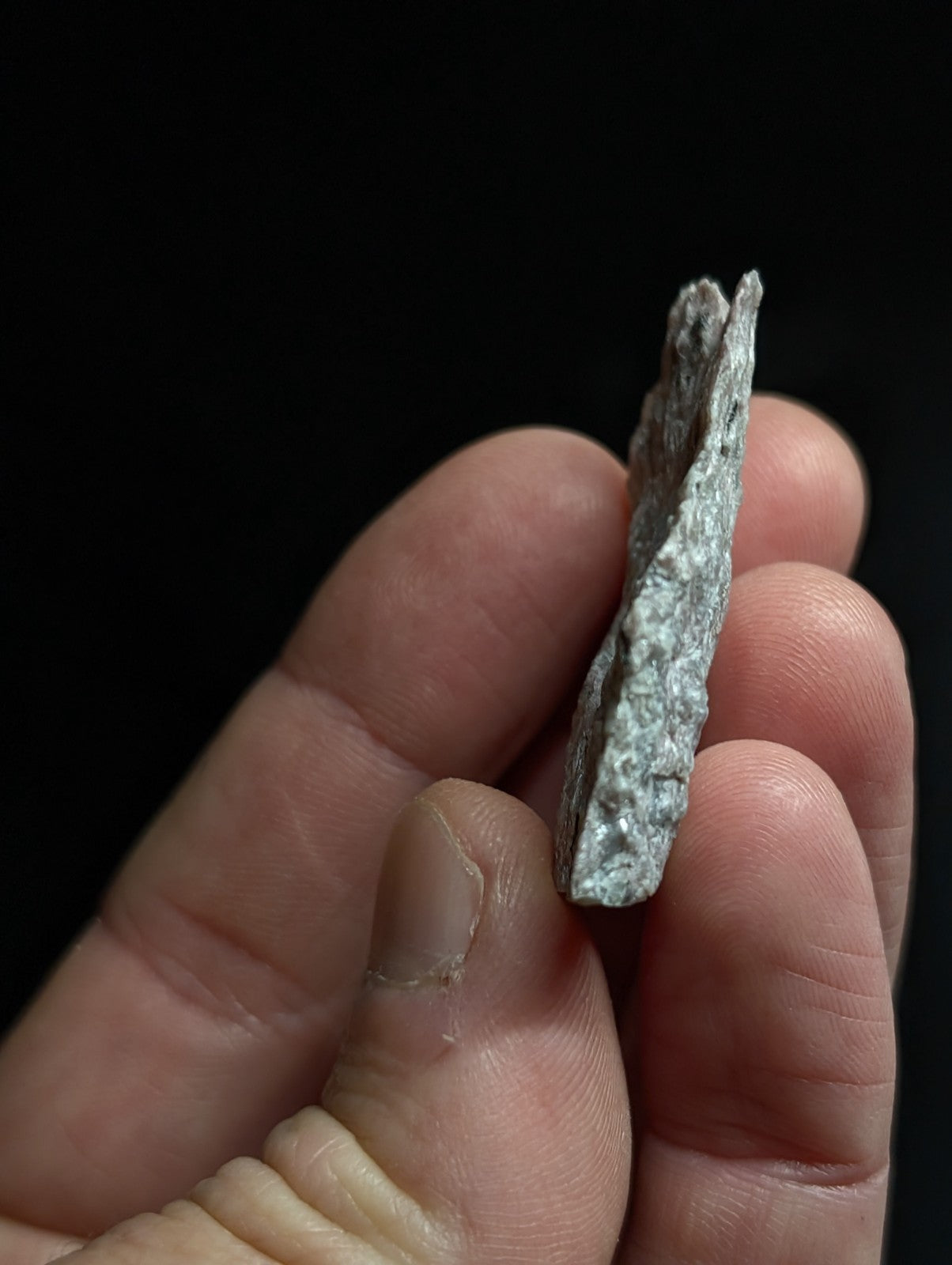 A weathered, grayish-green metallic fragment with a rough, jagged edge and uneven surface texture.