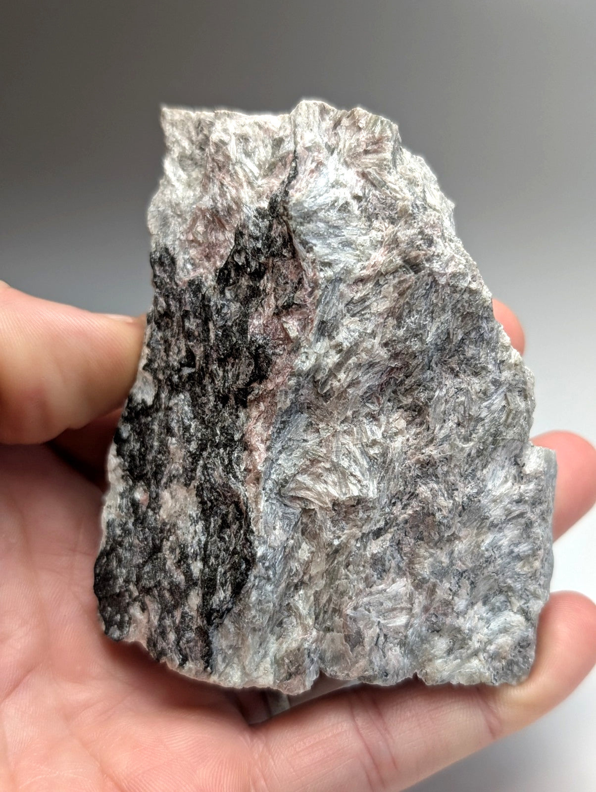 A hand holds a rough miserite and wollastonite mineral specimen with gray, black, and subtle pink hues from Arkansas