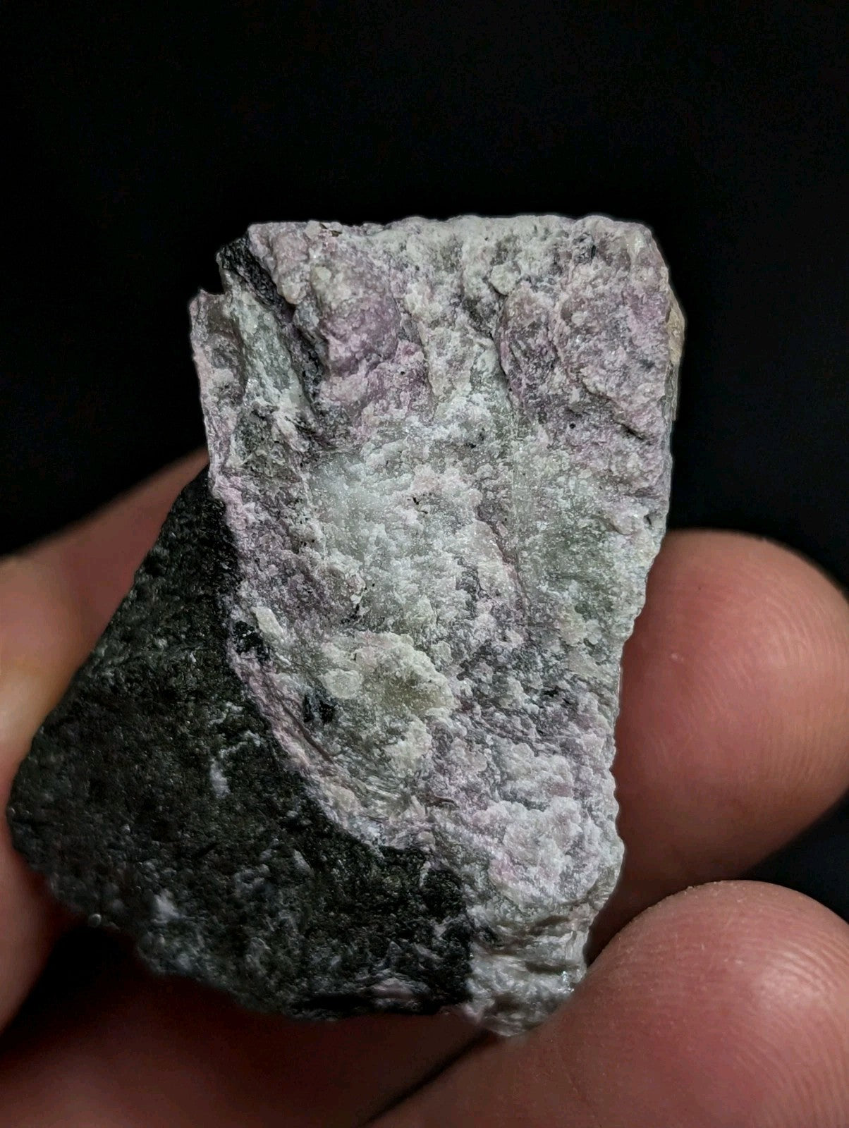 A rough, irregularly shaped mineral specimen with a mottled surface displaying patches of gray, white, and subtle purple hues.