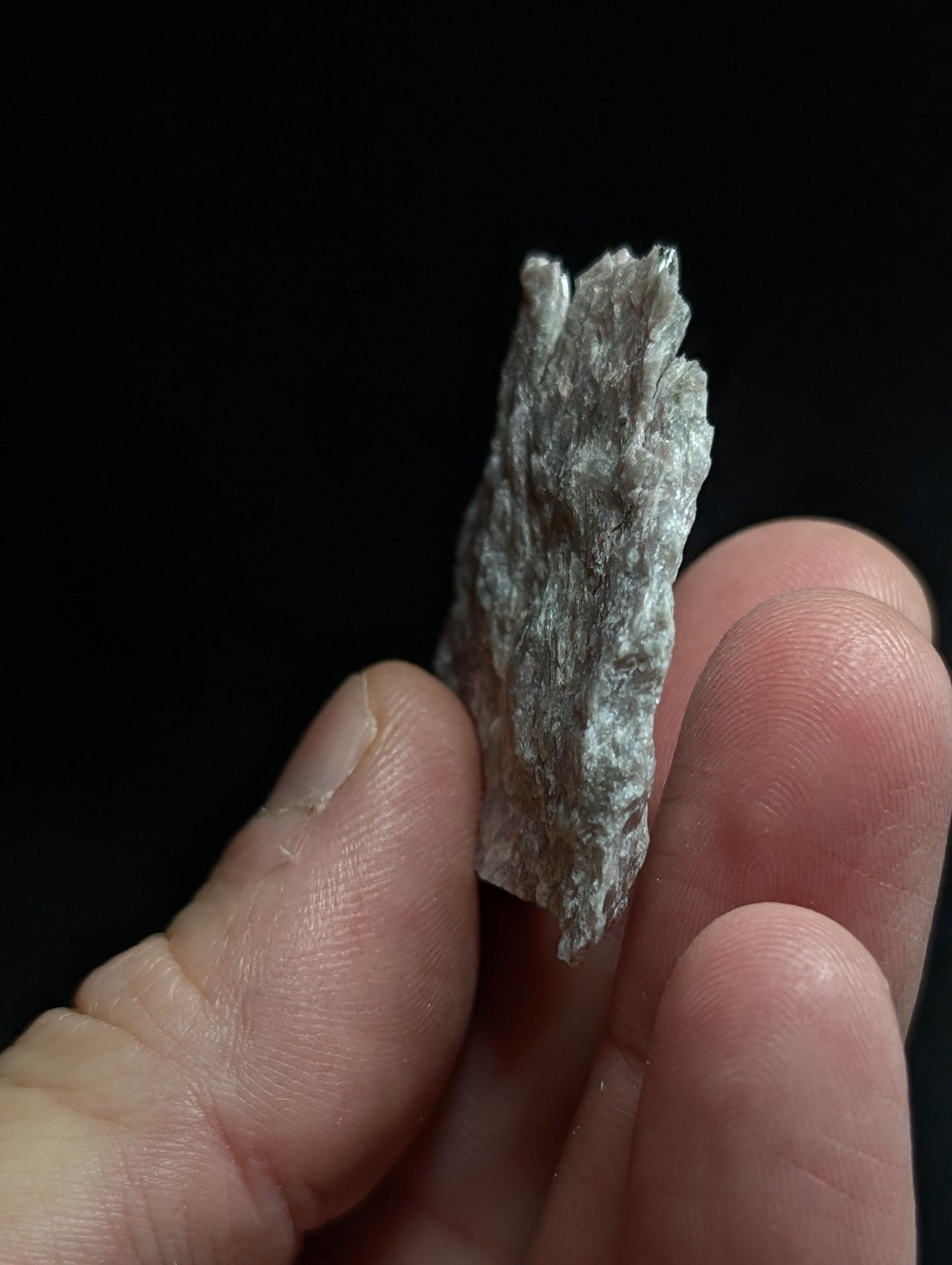 A rough, grayish-white mineral specimen with a textured, uneven surface held between fingers.