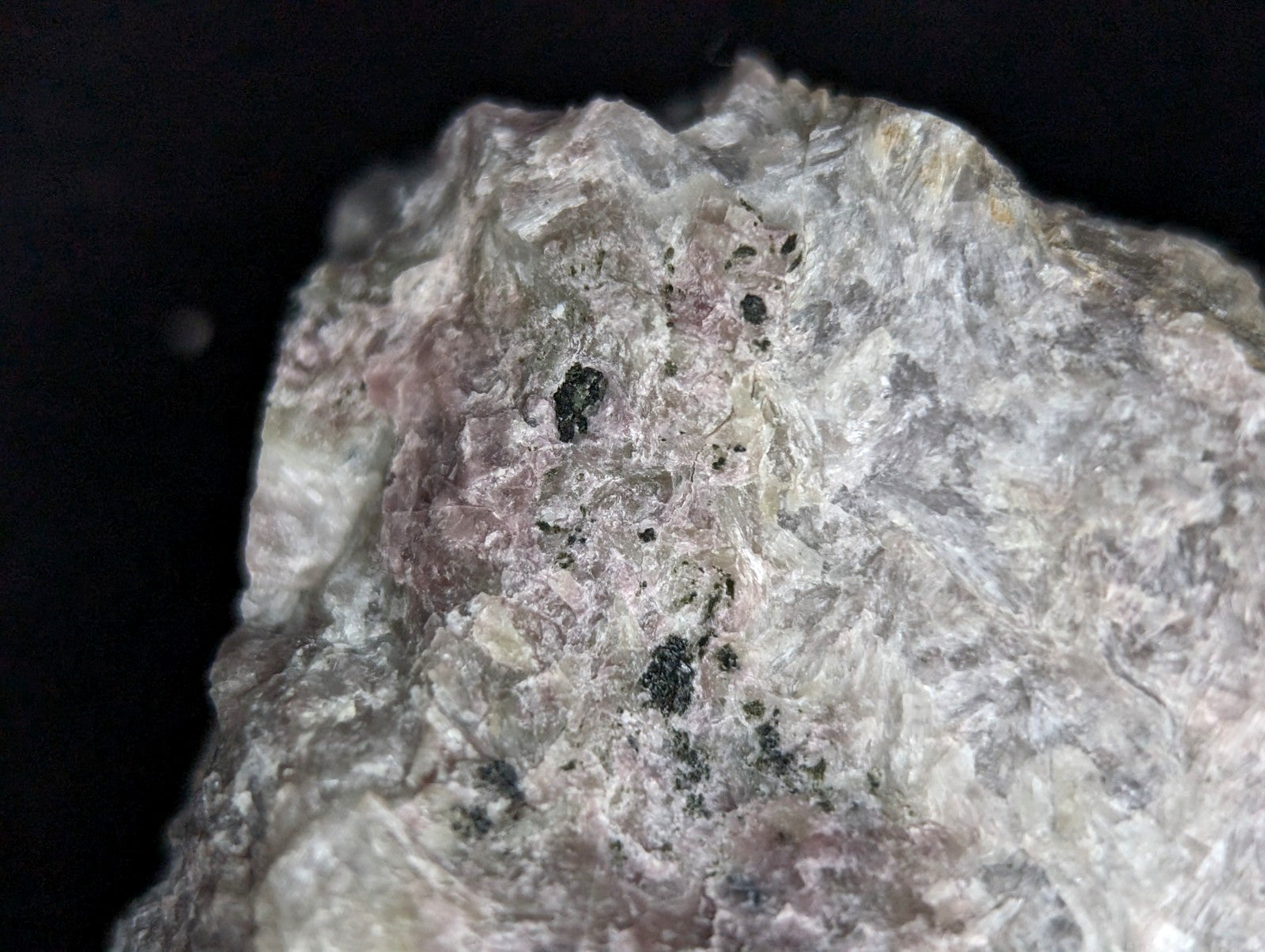 A rough, irregularly shaped rock with a mottled surface of white and pale pink hues, speckled with dark black inclusions.