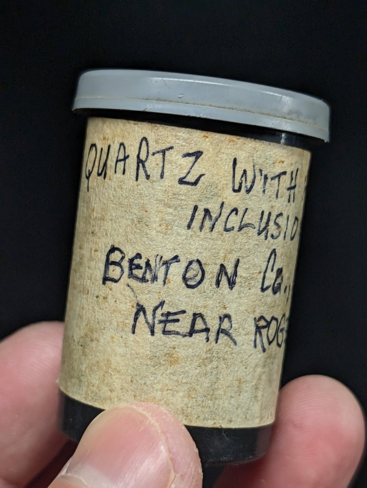 A vintage cylindrical mineral sample container with a gray lid and aged, handwritten label identifying its contents as quartz from benton co., near rogers.