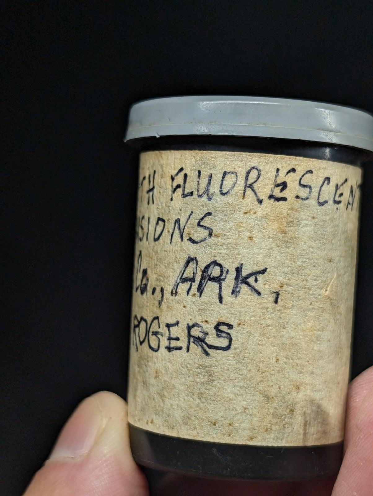A vintage cylindrical container with a gray lid and aged, handwritten label reading ’fluorescent’ and ’ark rogers’.