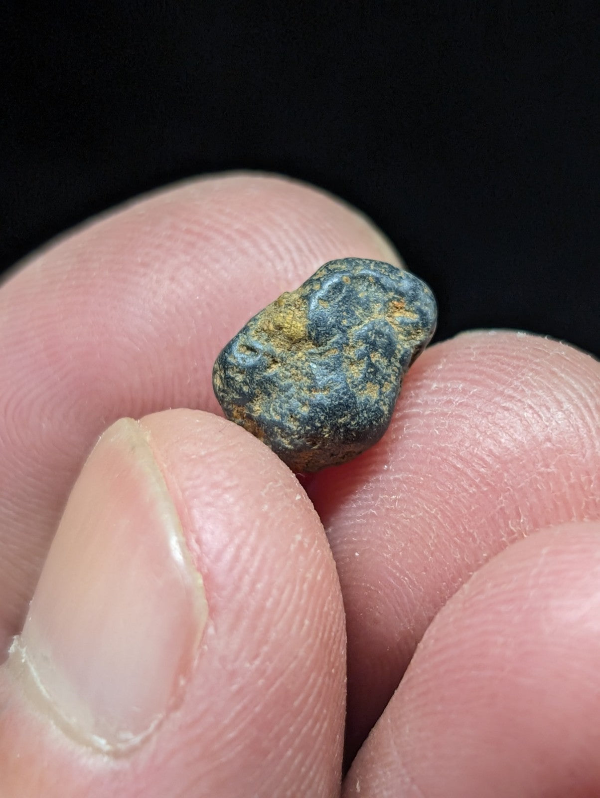 A small, irregularly shaped meteorite with a dark blue-gray surface speckled with patches of yellowish rust.