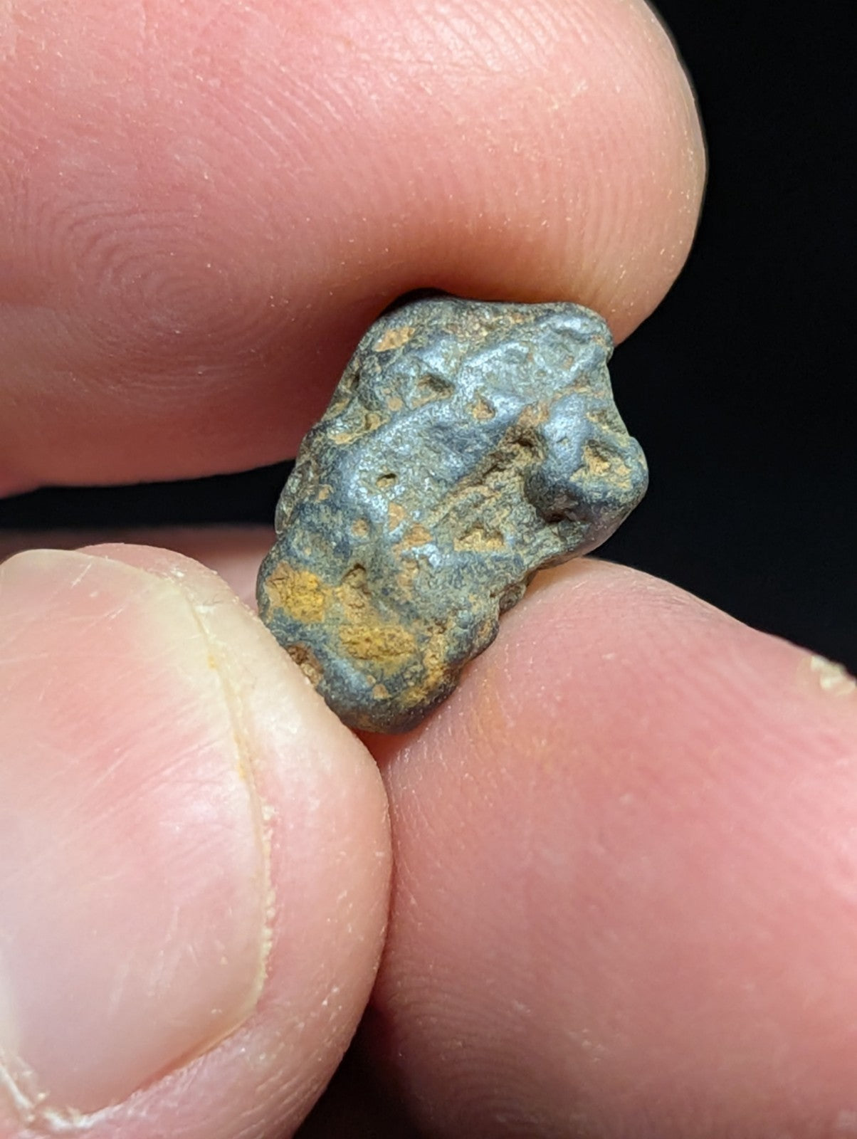 A small, irregularly shaped metallic object with a greenish-blue patina and patches of yellow oxidation, held between fingers.