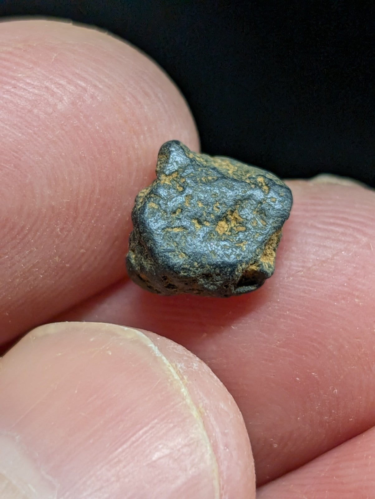 A small, irregularly shaped meteorite with a dark gray surface speckled with patches of yellowish rust.