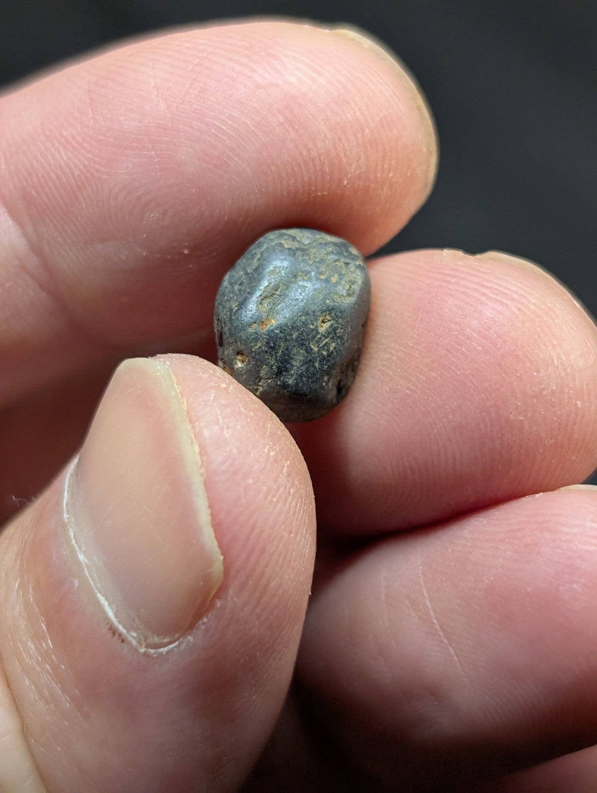 A small, dark gray, irregularly shaped stone with a rough, slightly pitted surface held between fingers.