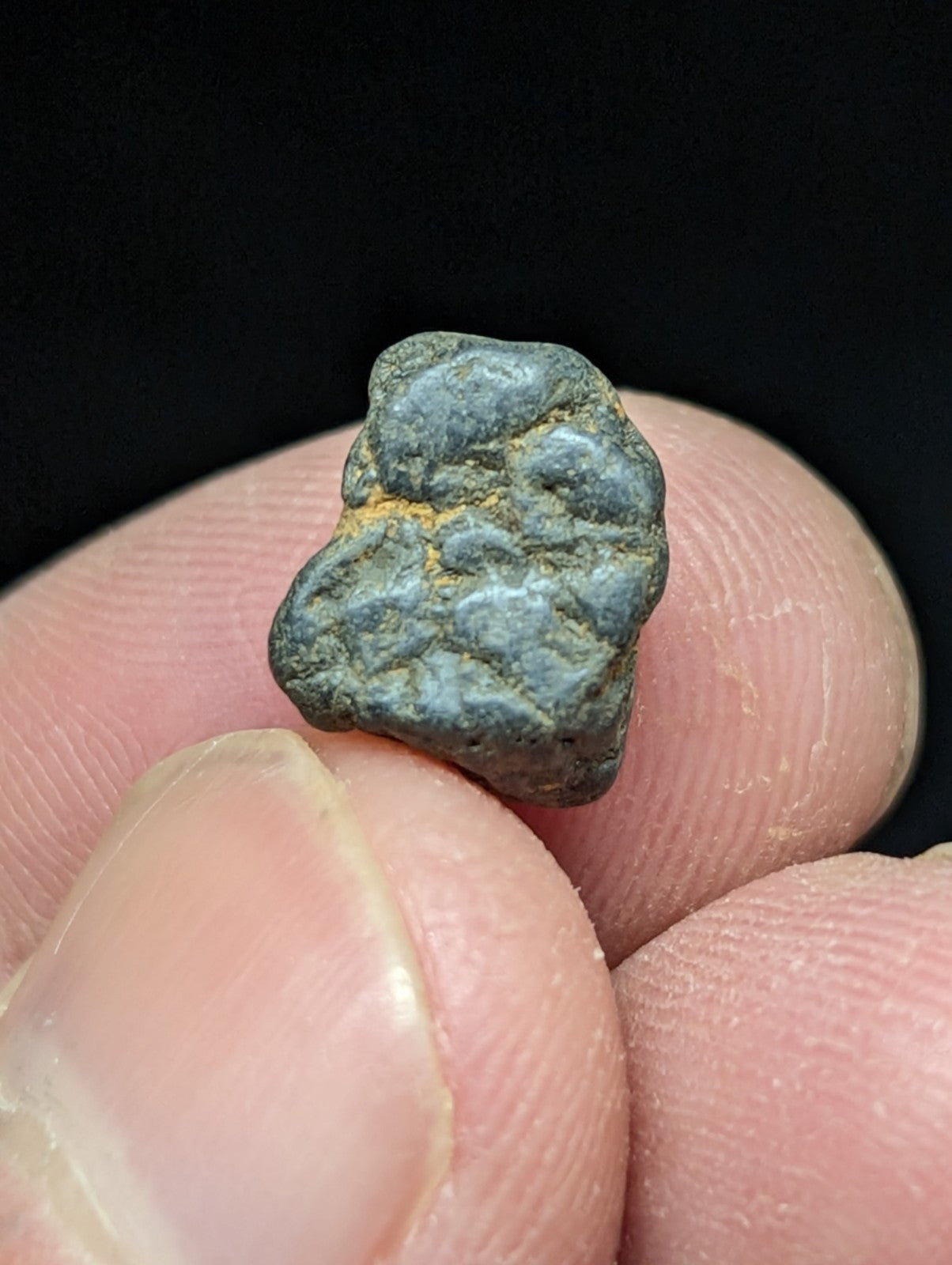 A small, irregularly shaped meteorite with a dark greenish-gray surface and patches of yellow oxidation.