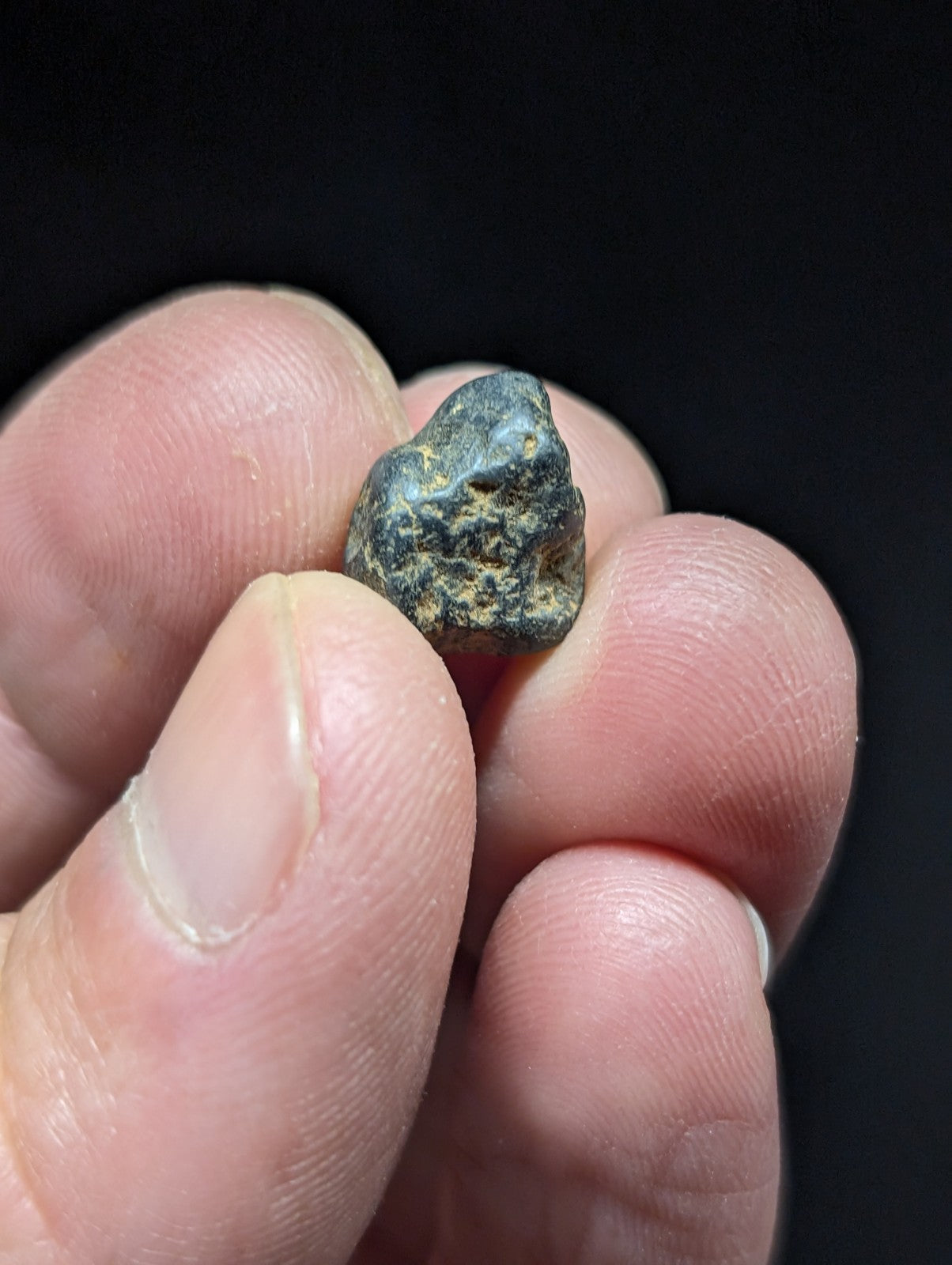 A dark, irregularly shaped rock with a rough texture and patches of lighter greenish-yellow mineral streaks.