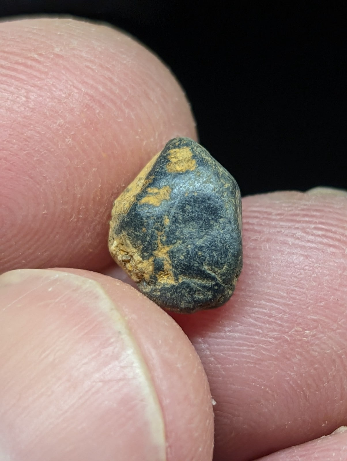 A dark, irregularly shaped stone with patches of yellowish mineral streaks held between fingers.