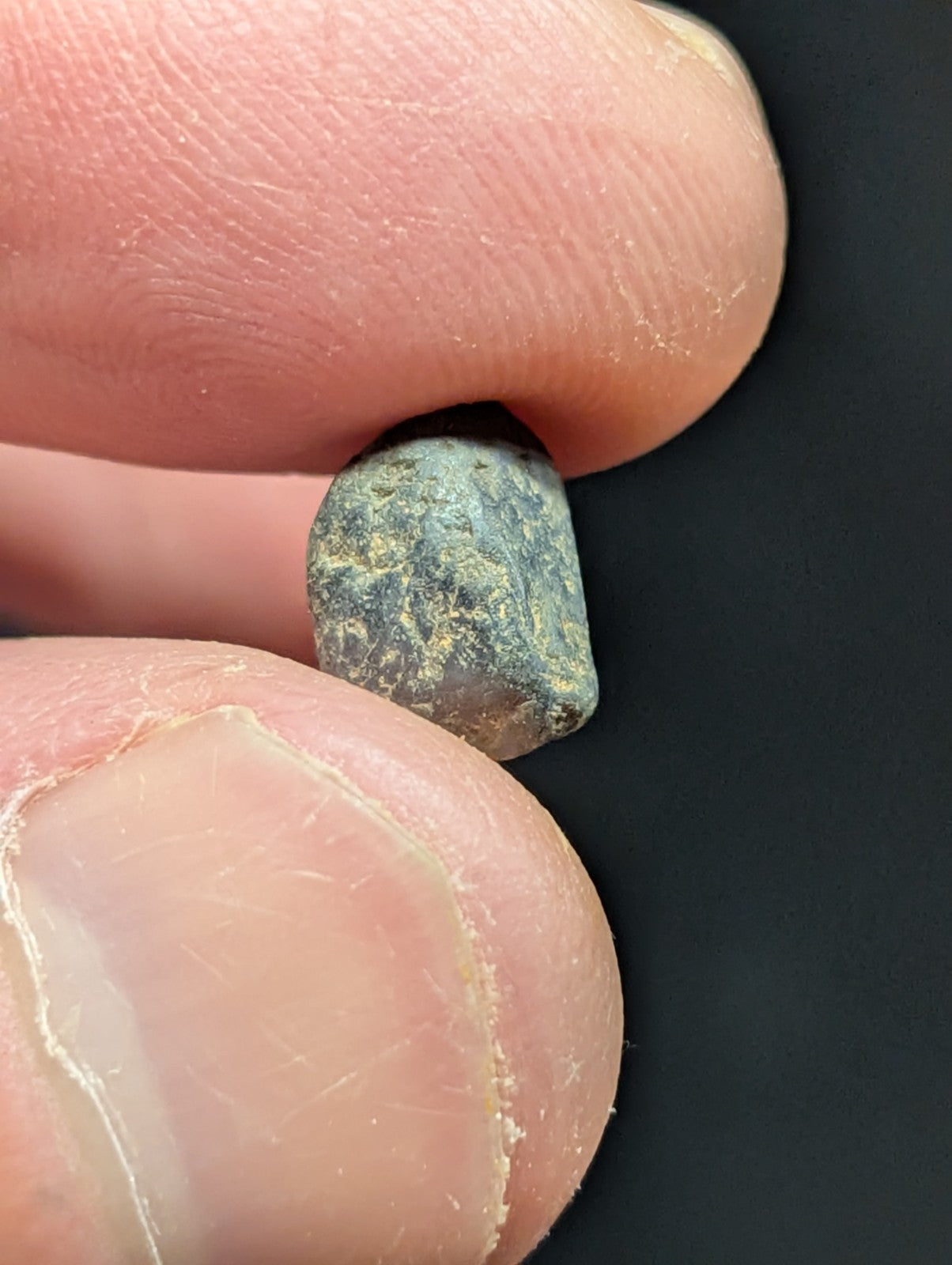 A small, irregularly shaped greenish-gray stone with a rough, textured surface held between fingers.