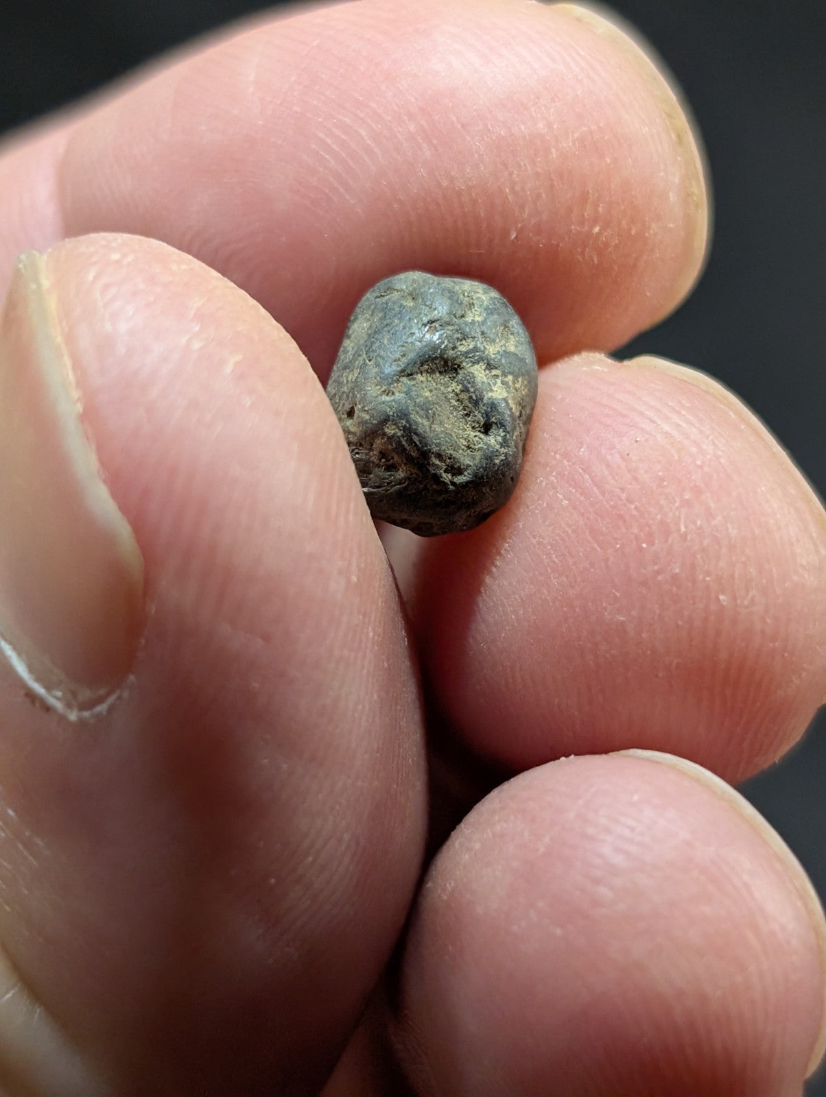 A small, dark greenish-gray, irregularly shaped stone with a rough texture held between fingers.