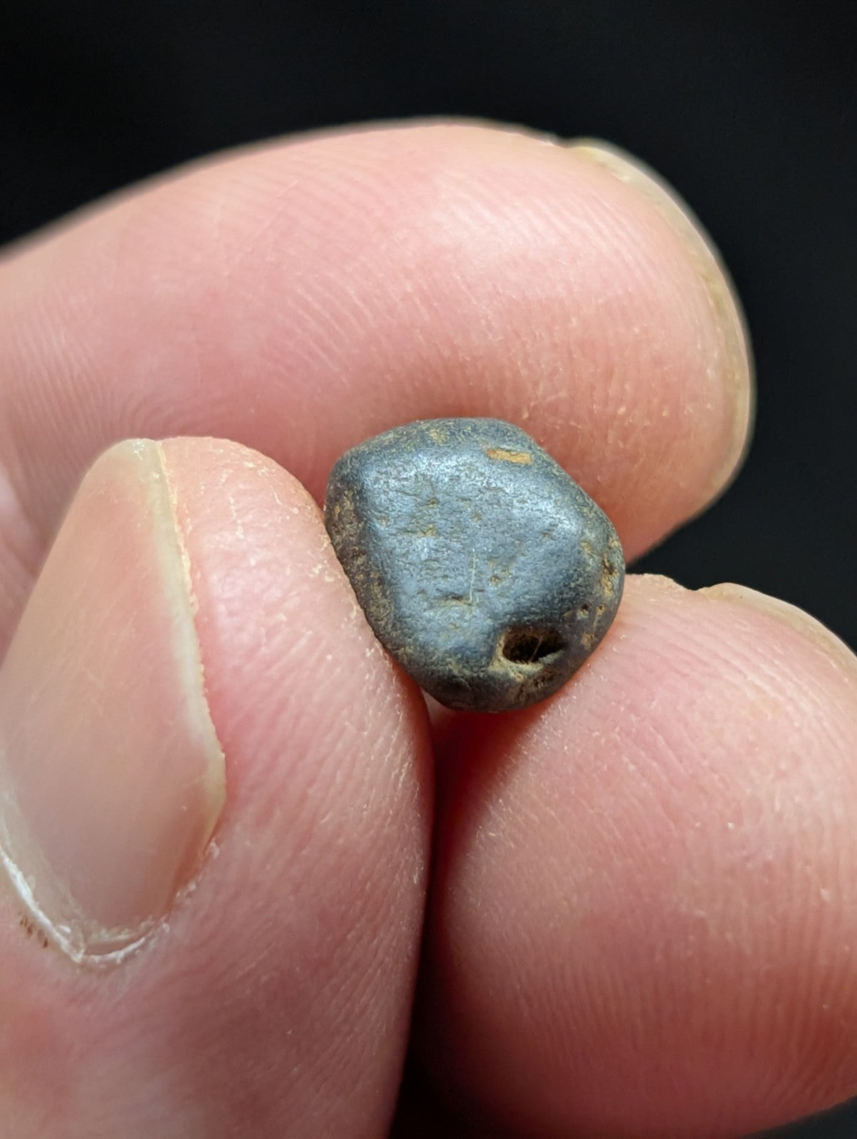 A small, dark gray, irregularly shaped stone with a rough texture and a noticeable hole near one end.