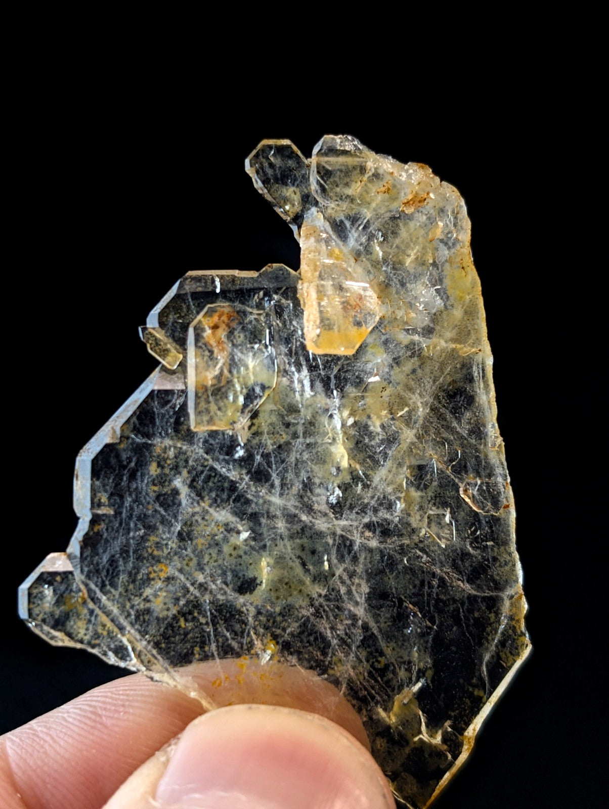 A translucent yellow-green mineral specimen with intricate crystalline formations and subtle orange inclusions, held delicately between fingers.