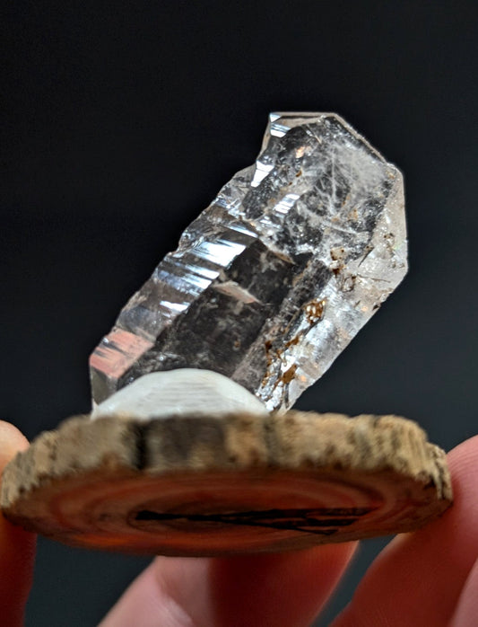 A clear, faceted quartz crystal with natural inclusions rests on a rustic wooden slice.