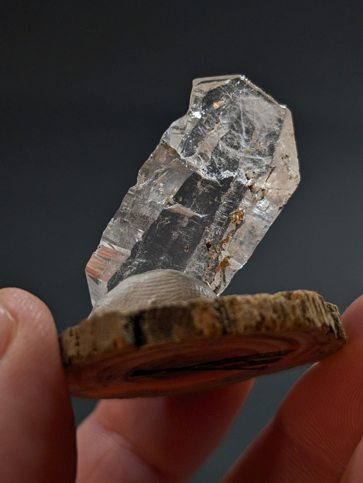 A clear, faceted quartz crystal with natural inclusions and a rough-hewn wooden base.
