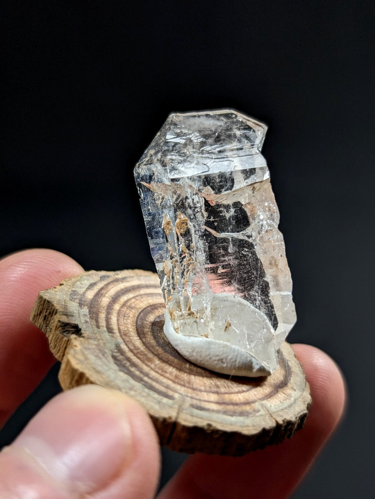 A clear, faceted quartz crystal with natural inclusions and a white base, displayed on a rustic wooden slice.
