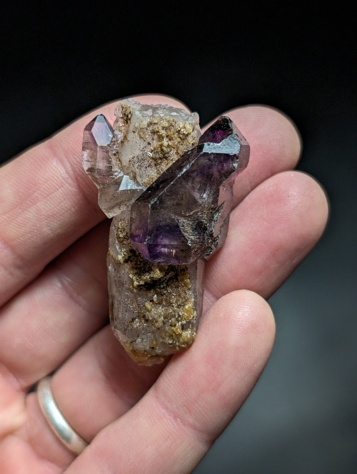 A hand holds a cluster of amethyst crystals with translucent purple and clear quartz formations, embedded in sandy matrix material.