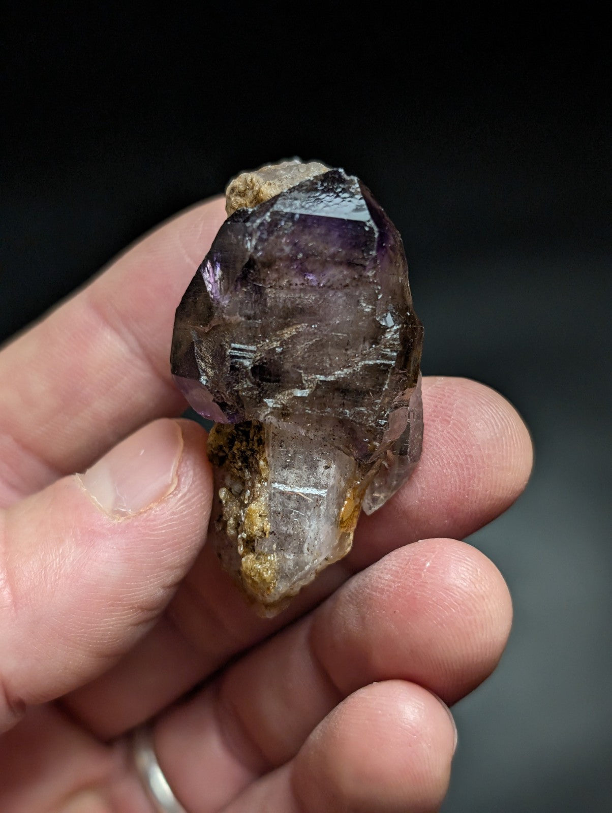 A hand holds a natural amethyst crystal cluster with translucent purple and clear sections, featuring earthy brown and yellow mineral inclusions.