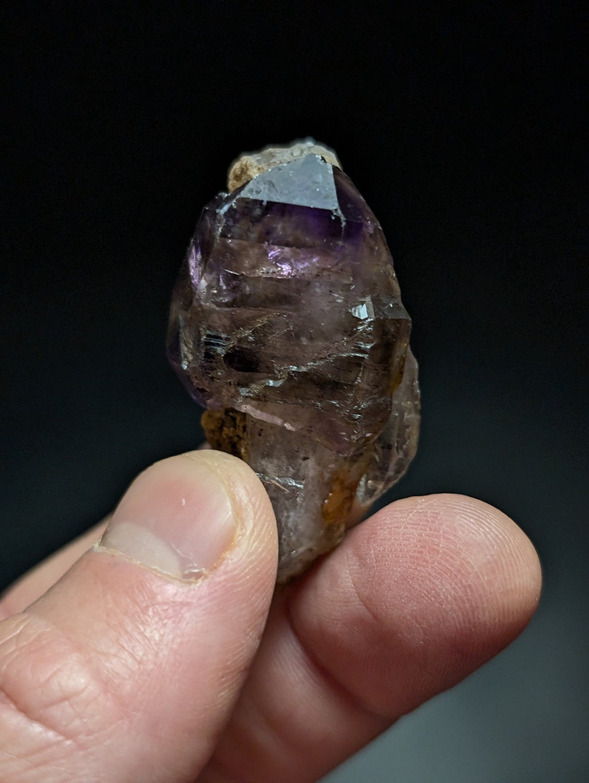 A translucent amethyst crystal with a pointed, faceted top and earthy brownish base held gently between fingers.