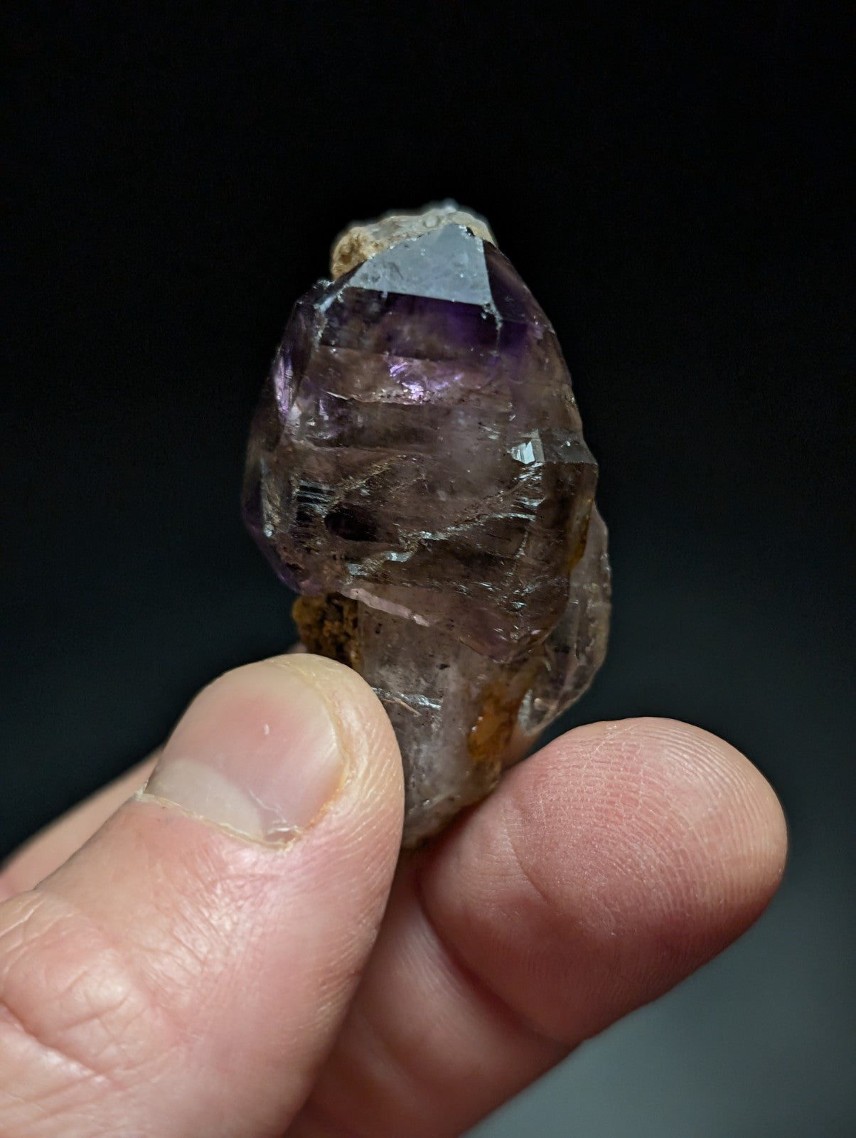 A translucent amethyst crystal with a pointed, faceted top and earthy brownish base held gently between fingers.