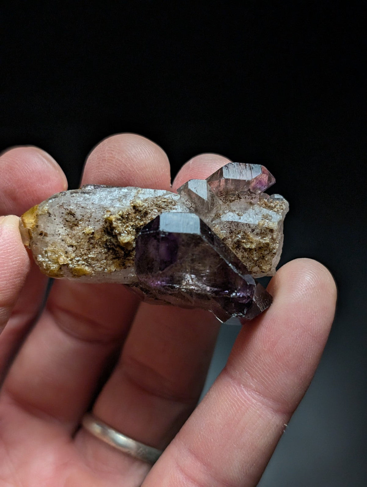A hand holds a natural quartz crystal cluster featuring translucent purple amethyst and clear, faceted crystals emerging from a rough, earthy matrix.