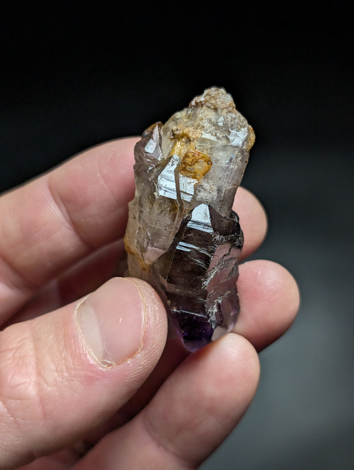 A hand holds a translucent amethyst crystal cluster with sharp, faceted points and natural brownish-yellow inclusions.