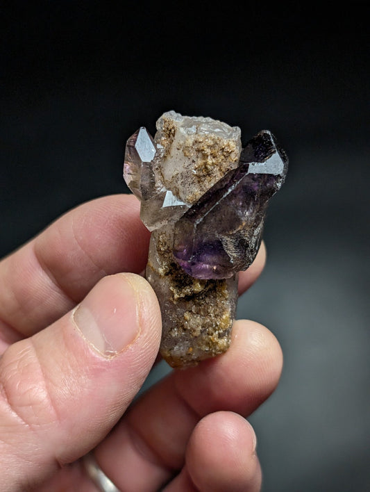 A hand holds a natural amethyst and quartz crystal cluster, featuring translucent purple and clear crystalline formations with earthy brown matrix.