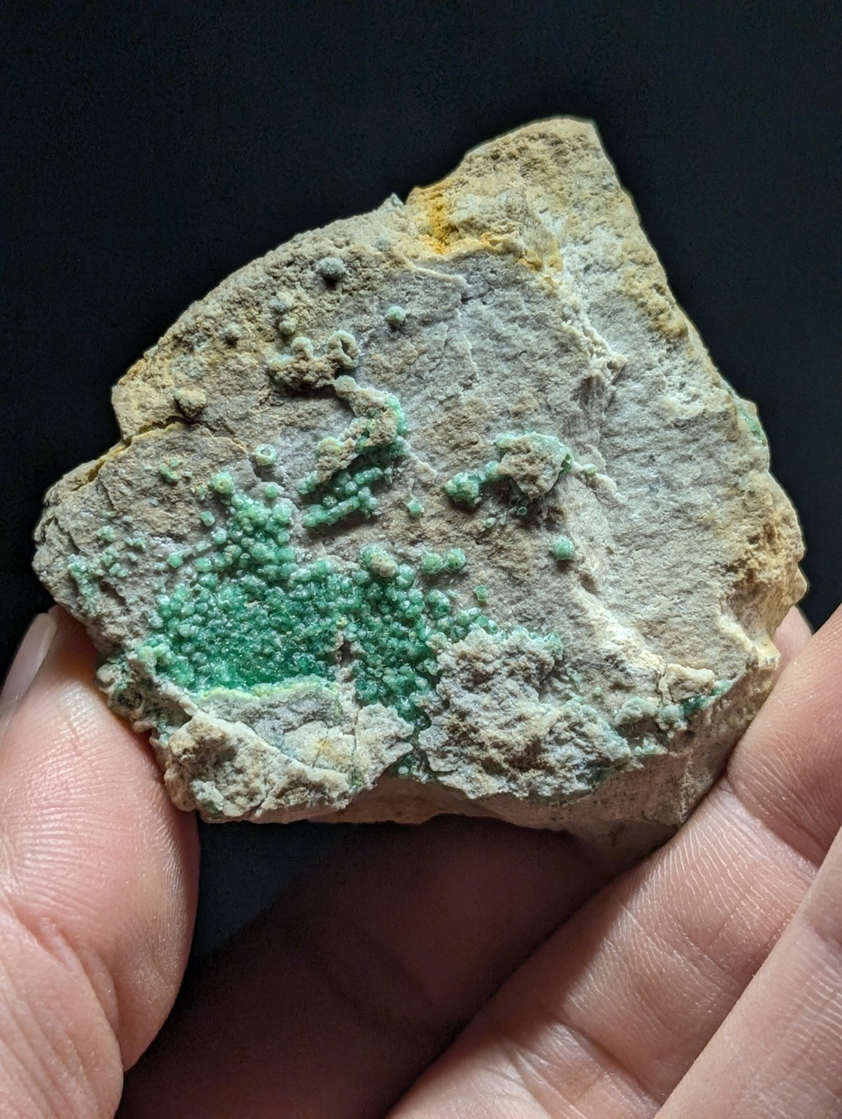 A hand holds a rough, irregularly shaped rock specimen featuring vibrant green crystalline formations against a pale gray and yellowish matrix.