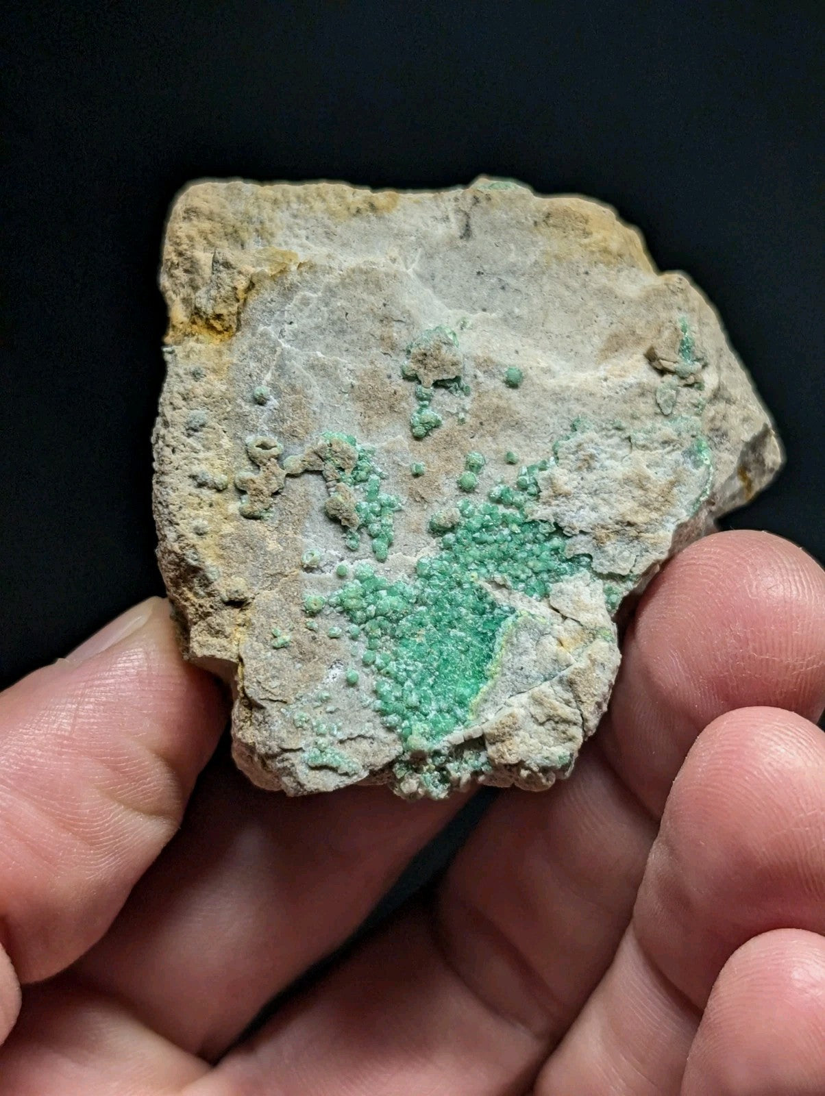 A hand holds a rough, irregularly shaped rock with patches of vibrant green mineral crystals embedded in its pale gray and yellowish matrix.