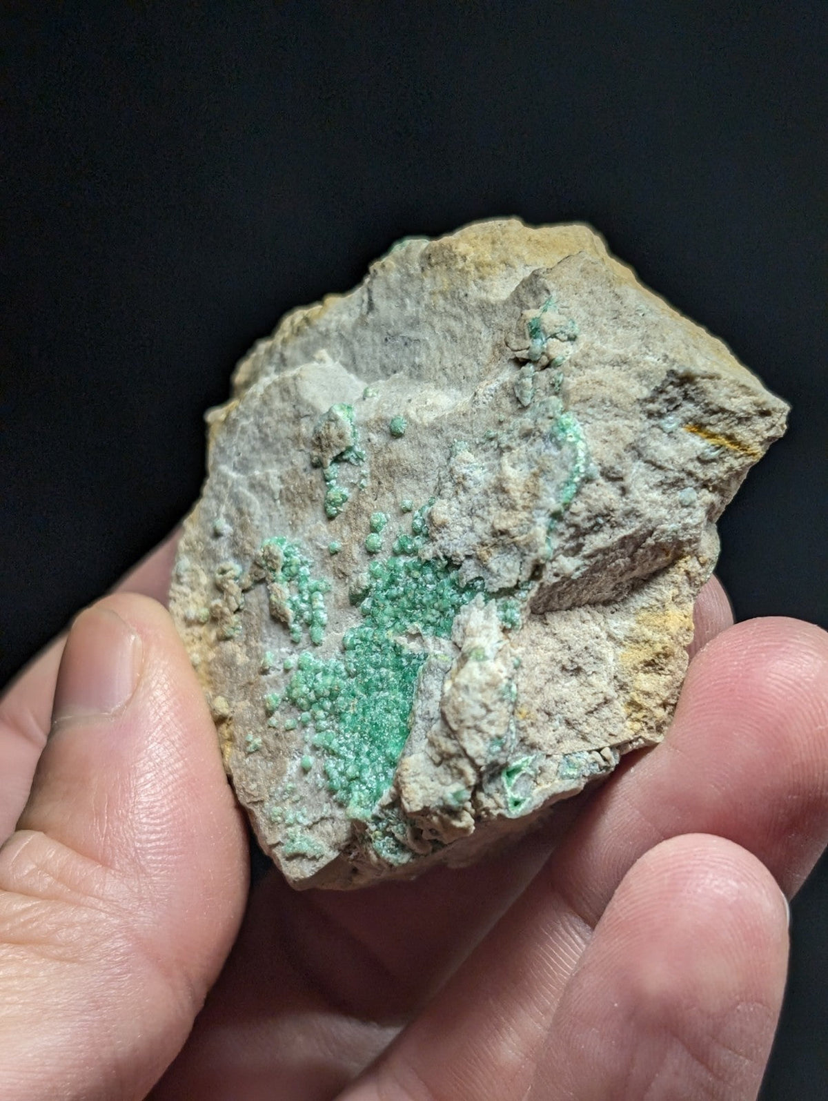 A hand holds a rough, irregularly shaped rock with patches of vibrant green mineral growth against a pale gray and yellowish matrix.