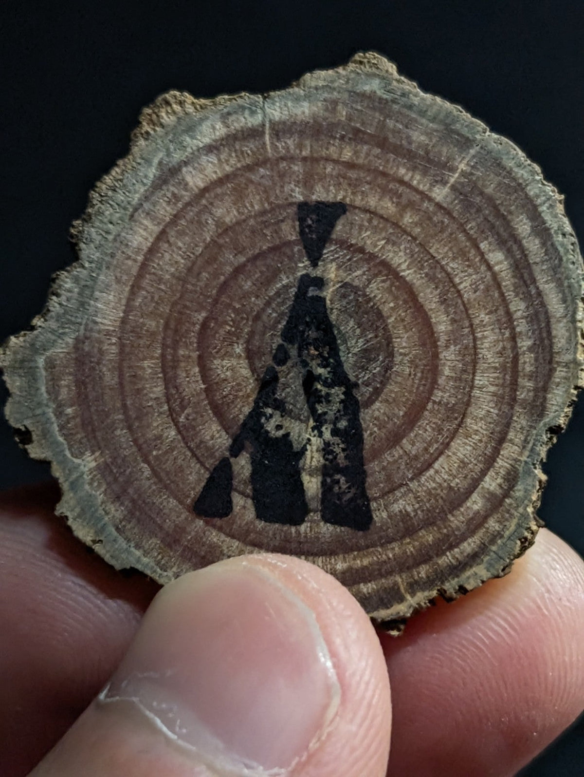 A circular wooden slice with natural grain and concentric rings, featuring a black painted silhouette of a figure.