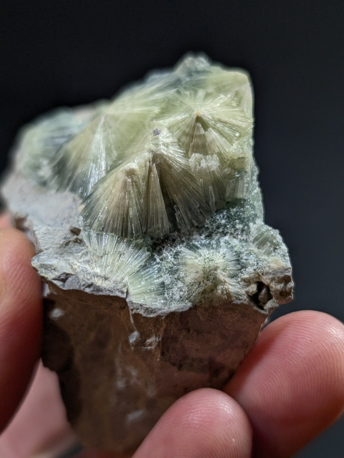 A hand holds a specimen of greenish-white, translucent calcite crystals with radiating star-like formations on a rough, earthy matrix.