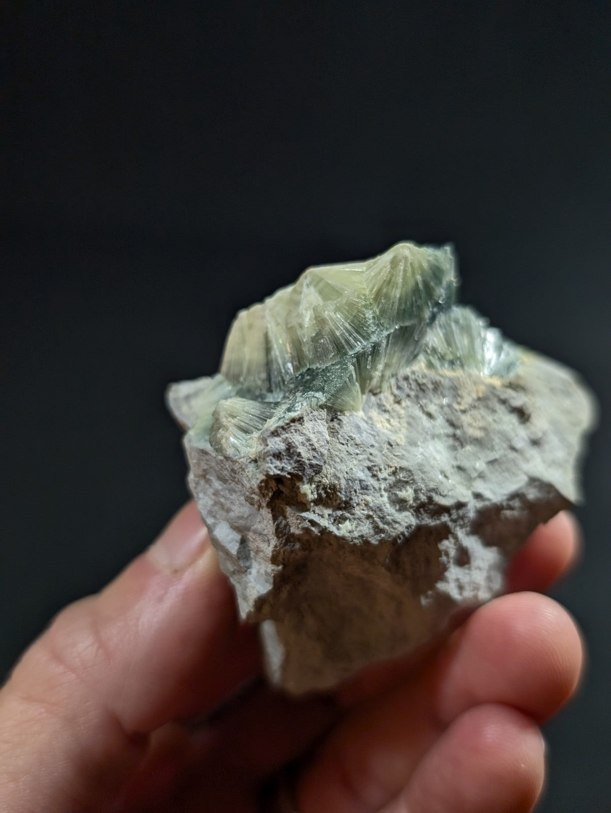 A hand holds a rough, irregularly shaped mineral specimen featuring translucent green crystalline formations atop a grayish matrix.