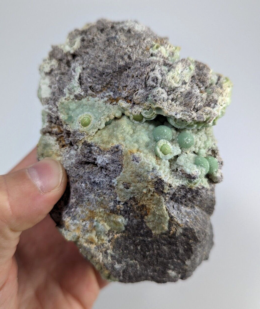 A hand holds a rough, irregularly shaped mineral specimen featuring grayish rock with patches of pale green and yellow-green crystalline formations that resemble small spheres or bubbles.