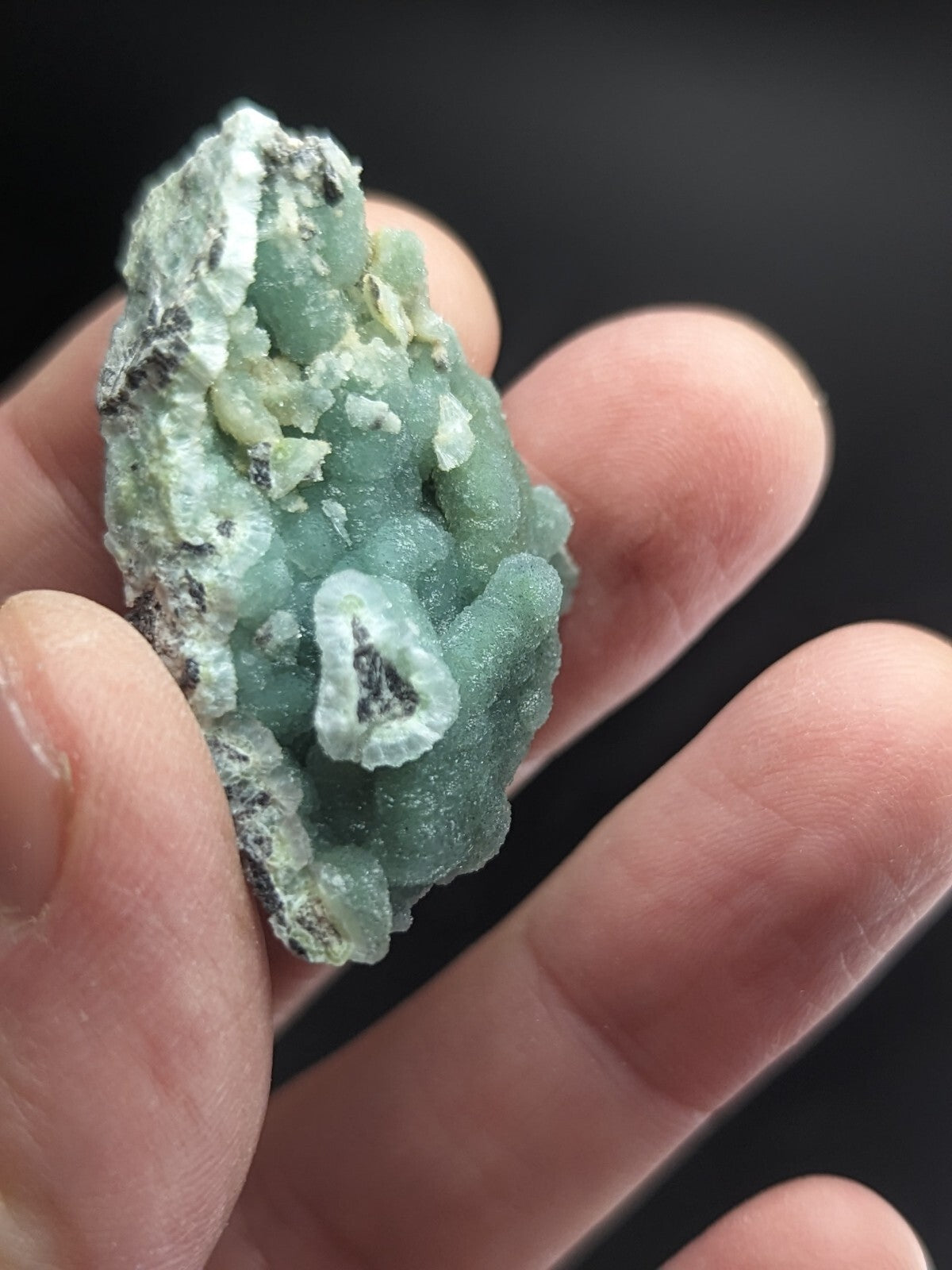 A hand holds a small, irregularly shaped specimen of greenish-blue mineral with white and gray crystalline formations.