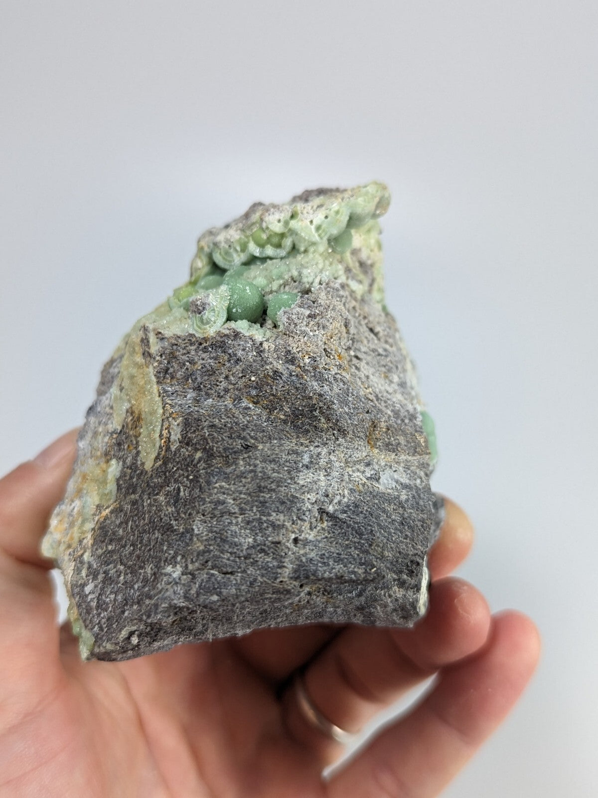 A hand holds a rough, gray rock specimen featuring embedded light green mineral formations.