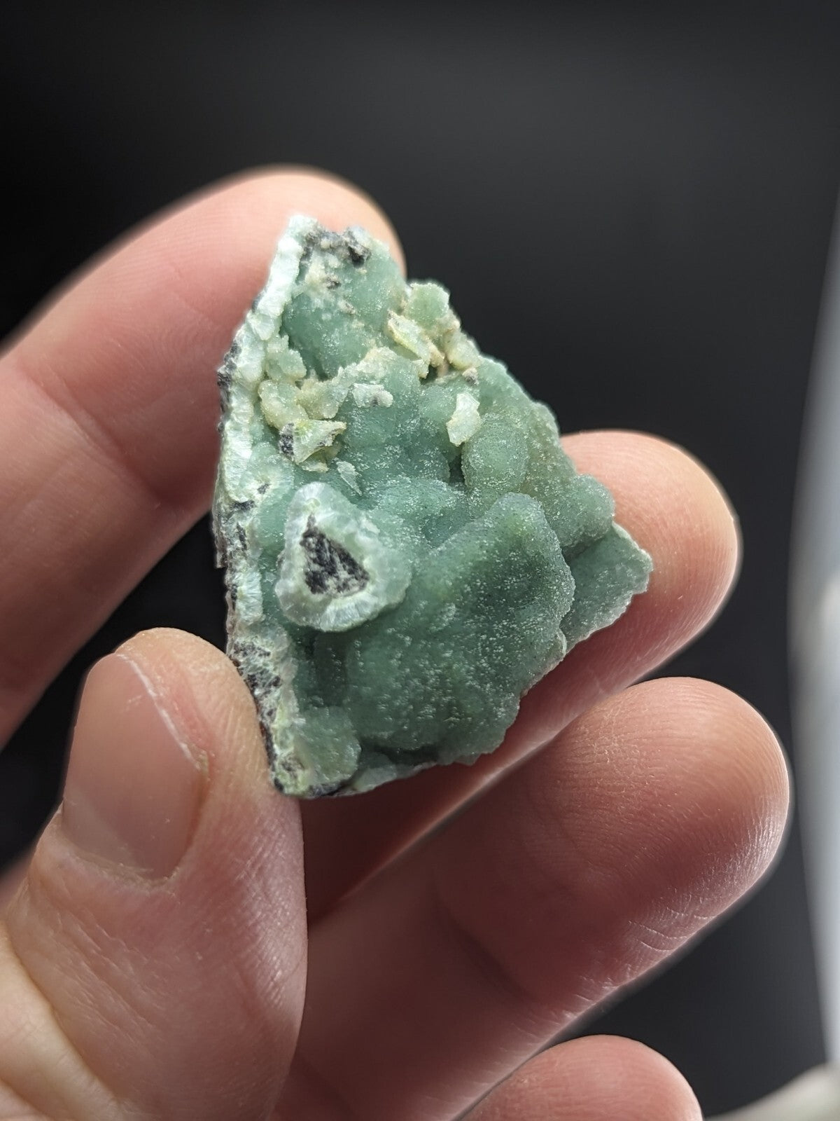 A hand holds a small, irregularly shaped green mineral specimen with a textured surface and white crystalline inclusions.